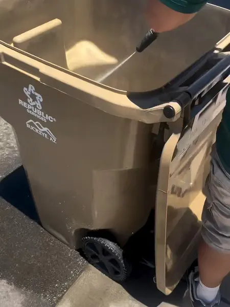 Hot-water pressure washing the inside of a tan trash bin at the curb