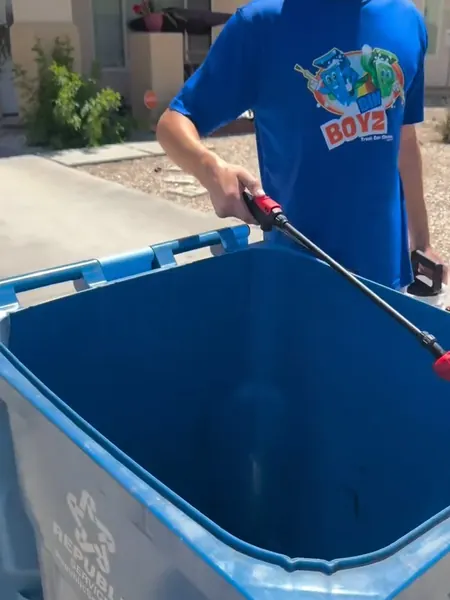A Bin Boyz crew member in a Bin Boyz t-shirt pressure-washing a blue recycling bin at the curb