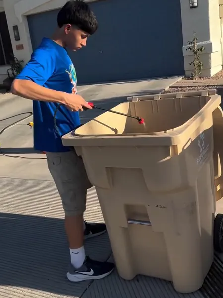 A Bin Boyz crew member washing a tan trash bin in a Buckeye driveway